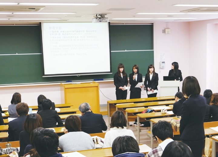卒業研究発表を行っている３人の女子学生と聴衆の学生、教員